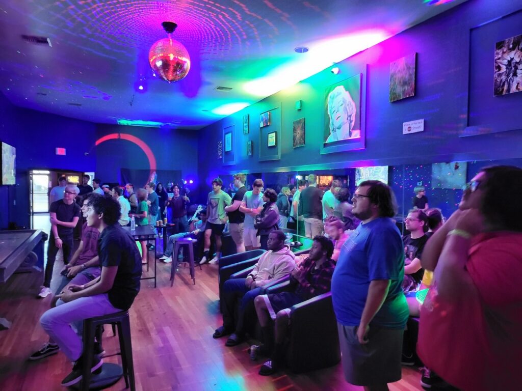 A crowd playing console games under a disco ball at a Play Money party